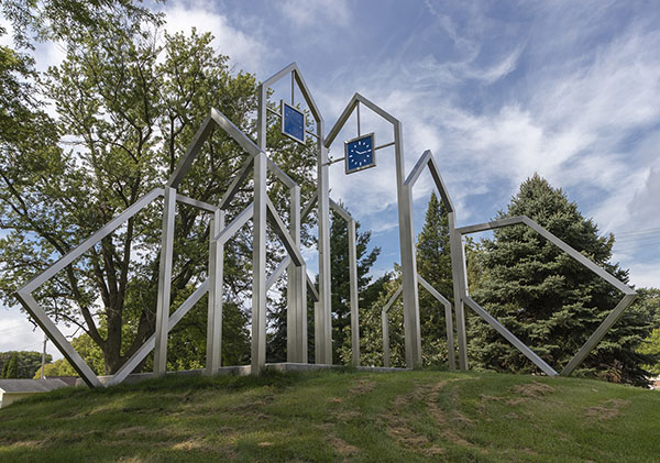 Westside Rising Flood memorial, pictured in 2018 before the storm. Steel outlines of houses, with a few tipped over, and the two tallest ones have blue clocks set to 10:15. There are evergreens and deciduous trees all around it.