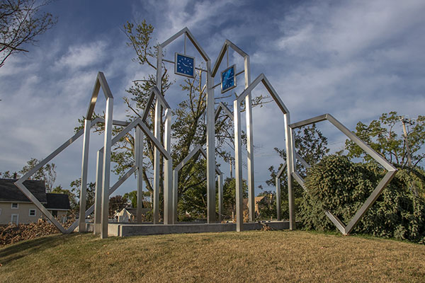 Westside Rising Flood Memorial in Time Check, Cedar Rapids: steel sculpture of the outlines of eight houses; two are tipped over, while the two tallest ones have blue clocks set to 10:15; a felled evergreen torpedoes through one of the tipped over houses