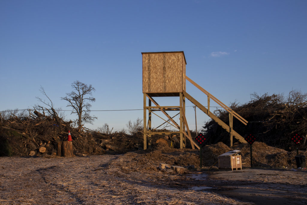 A wooden structure that resembles a hunting shelter, with wooden steps leading up to a small, square building made of wood. It stands in the center of a clearing, surrounded by tree limbs and logs, and next to the stairs is a crate that is partially open. On the other side, an emergency cone is balanced atop a log.