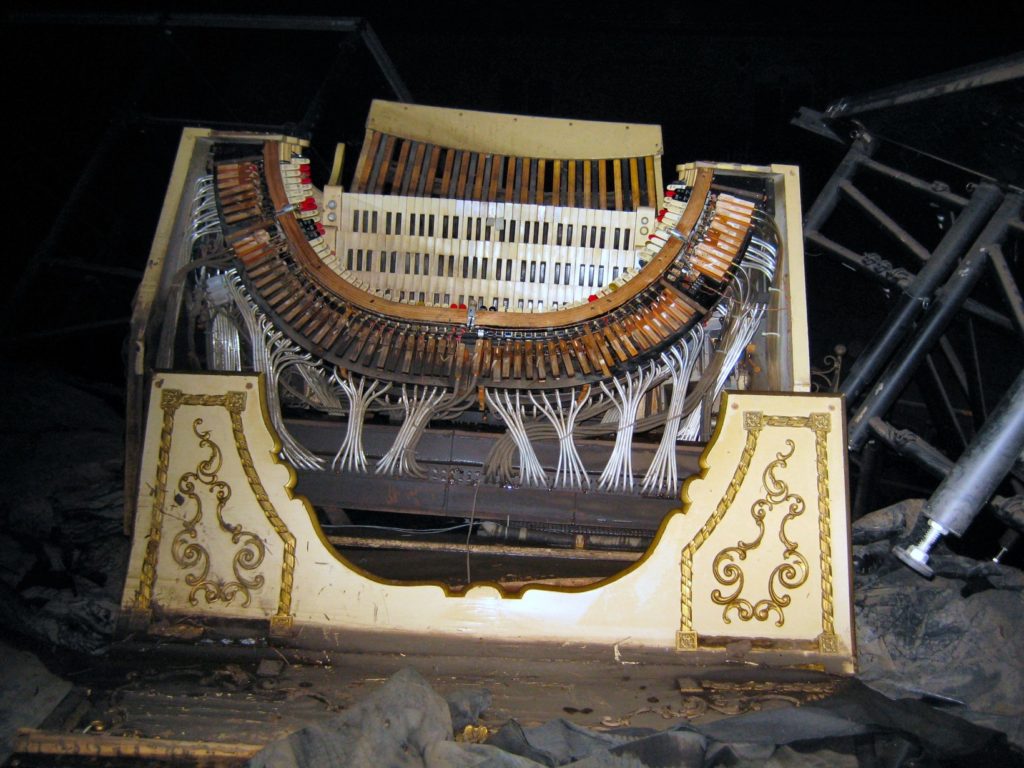A color photo of the Mighty Wurlitzer organ toppled over, in a dark theater with no lights except the photo lights aimed at the organ. It has extensive damage. Photo credit: Cedar Rapids Gazette